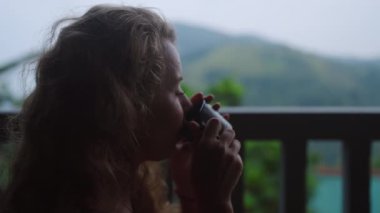 Günlük rahatlık, sabah sükuneti, doğa arkaplanlı ferahlama. Kadın çayını yudumluyor salıncakta dinleniyor, lüks dağ manzaralı teras çay bitkilerini görüyor. Rahatlık, yalnızlık bitkisel içecekle.