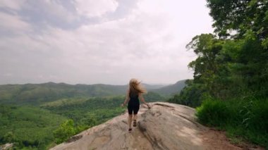 Kaya üzerinde duruyor, kolları açık, özgürlük hissi, seyahat yaşam tarzı. Kadın dağ zirvesine tırmanıyor, geniş yeşil manzaranın tadını çıkarıyor. Tek başına doğa yürüyüşü, macera, refah, temiz hava soluyor. Slomo