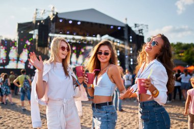 Bir grup genç kadın kutlama yapıyor, açık hava konserinde eğleniyor, kalabalıkla dolu canlı bir festival sahnesi, arka planda sahne. Kızlar plajda yaz müzik festivalinin keyfini çıkarıyor, içkilerini yudumluyor, gün ışığında dans ediyor..