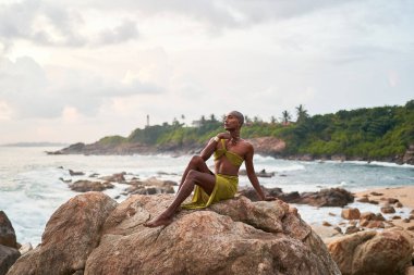 Androgynous siyah model gün batımında okyanus manzaralı sahilde poz veriyor. Uzun ve gösterişli bir elbiseyle ikili olmayan etnik model gün doğumuna karşı deniz kenarında kayalıklarda oturur. Okyanus arka planında karanlık. LGBTQ