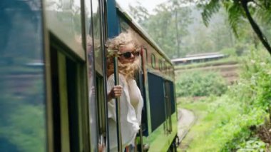 Kadın turist manzaralı tren yolculuğunun keyfini çıkarıyor, yeşillik dolu bölgede tatilde özgürlüğünü hissediyor. Maceraperest kadın tren kapısından dışarı sarkmış, sarı saçlı rüzgar, verimli tropikal manzara geçip gidiyor. Yavaşla