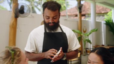 Bipoc dostu önlüklü garson tropikal restorandaki bayan müşterilerden sipariş alıyor. Kafe çalışanı kafe menüsünü tavsiye ediyor. Çekici garson konuşuyor, yemek seçiminde misafirlere yardım ediyor..