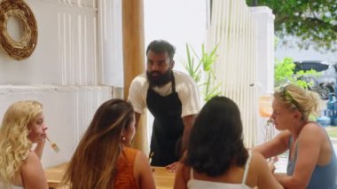 Siyah garson Bali 'deki kafe terasında bayan müşterilerle konuşuyor. Çeşitli genç kadınlar yemek yer, açık hava restoranındaki kafe işçisiyle menüyü tartışır. Dost canlısı garson müşterilerine menü öneriyor.