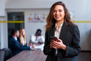 Profesyonel kadın mailleri kontrol eder, görevleri yönetir, kameraya gülümser. Kendine güveni tam, sarışın iş kadını şık giyinmiş. Ofiste akıllı telefon kullanıyor. İş arkadaşları arka planda buluşuyor..