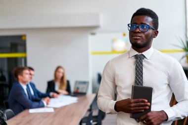Meslektaşlar arka planda iş tartışır. Modern ofisteki kendinden emin Afrikalı işadamı, tableti elinde tutuyor, sunuma hazır. Pro, çeşitli işyerlerinde liderlik, iş dünyasında teknoloji.