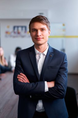 Toplantıda arka planda çalışan meslektaşlar. Kendine güvenen genç işadamı kollarını kavuşturup gülümsüyor. Profesyonel erkek mavi takım giyiyor, beyaz gömlek giyiyor, kravatı yok, liderlik örneği gösteriyor..