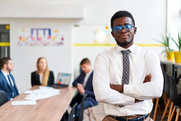 Teamwork in background, inclusive and diverse work environment. Confident African businessman stands in modern office, arms crossed. Pro attire, young leader, business strategy session.