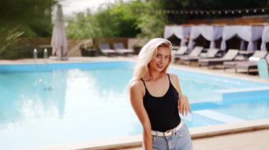Sarı saçlı kadın kırmızı güneş gözlüğü, şort ve yüzme havuzunda güneş pansiyonları ve çam ağaçlarıyla fotoğraf çekmek için dans ediyor. Tatillerde poz vermek ve flört etmek.