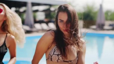 Uzun saçlı, dans eden ve plaj kulübünde açık bikinili yüzme havuzunun yanında flört eden kadın modeller. Yaz tatili için güneş gözlüklü kızlar poz veriyor ve kameraya bakıyor. Yakın plan..