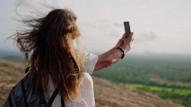 Canlı ruh, deneyimli yürüyüşçü yüksek irtifada maceradan hoşlanıyor, anıları kaydediyor. Enerjik kıdemli Asyalı kadın dağ zirvesine ulaşır, rüzgar akıllı telefonda nefes kesen bir manzara gibi saçları okşar. Yavaşla