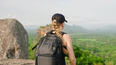 Duraklamalar, telefonlar, manzara manzarası, kayıt seyahati. Sırt çantalı bir kadın dağdan aşağı iniyor. Arka planda Sigiriya Kayası var. Gezgin araştırıyor, tarihi Sri Lanka simgesini belgeliyor. Yavaşla