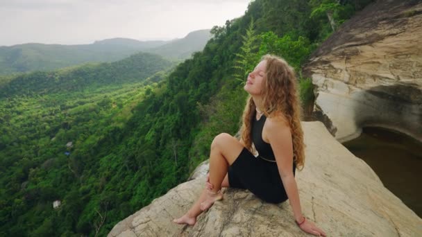 她放松下来 沉浸在宁静的自然景观中 感到自由 在富饶的绿色丛林里 卷曲头发的女人坐在悬崖边俯瞰瀑布 历险旅客享有生态旅游目的地 — 图库视频影像
