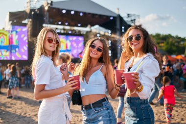 Günlük plaj kıyafeti, güneş gözlüğü, sahne arkası. Üç kadın, güneşli bir sahil müzik festivalinin tadını çıkarıyor. İçkilerini yudumluyor, gülüyor, dans ediyor. Millennial yaz partisi havası.