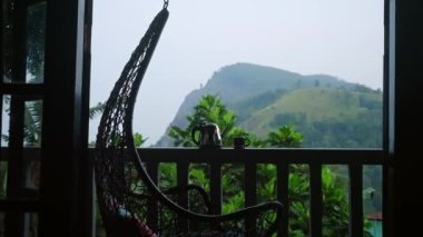 Teras korkuluklarında çaydanlık ve bardak, görülebilir çay tarlaları olan panoramik dağ manzarası, sisli sabah atmosferi, sallanan sandalye hafifçe doğaya lüks bir kaçış yapıyor. Yavaş çekim.