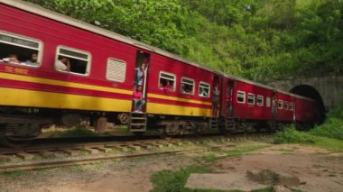 2023-06-01 Ella, Sri Lanka. Dokuz Arch Köprüsü. Kırmızı tren tünelden viyadük 'e çıkıyor. Yolcular yeşil bitkiler arasında eğilir, el sallar, anları yakalar.