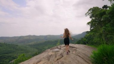 Kolları geniş, saf özgürlüğü hissediyor, doğanın zirvesinde neşe. Siyahlı kadın dağa tırmanıyor, geniş yeşil manzarayı kucaklıyor. Tek başına gezen gezgin, ferahlatıcı manzaranın, vahşi doğanın huzurunun tadını çıkarıyor. Slomo
