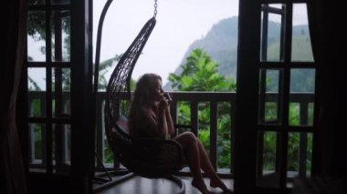 Kadın, Ella, Sri Lanka 'da huzur dolu lüks bir seyahatin ortasında sakin dağ manzaralı, lüks bir terasta sallanan bir salıncak sandalyesinde sabahın erken saatlerinde çaydan hoşlanıyor. Yavaş çekim.