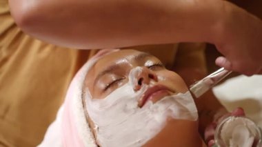 Kozmetik uzmanı, kadın hasta yüzüne sağlık merkezinde bir fırçayla beyaz kilden maske takıyor. Güzellik uzmanı, kaplıca çekiminde genç bir bayana yüz bakımı prosedürü uyguluyor. Güzellik ve yüz bakımı kavramı