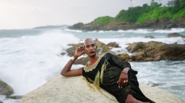 Lüks elbiseli, el yapımı mücevherleri dalgalı deniz kıyısının üzerinde duran tuhaf siyahi bir adam. Ağır çekim. Lgbtq iki ırklı moda eşcinsel modeli, şık bir elbise, pirinç aksesuar portresi. Tropik konum.