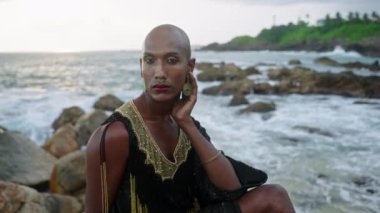 Lüks elbiseli eşcinsel bipoc adam, butik pirinç mücevher, tropik kayalık okyanus sahilinde otururken makyaj pozları. Homoseksüel epatage mankeni, gösterişli bir elbise, piercing, küpeler, mücevherli yüzükler..