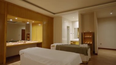 Geniş bir spa odasının ortasında masaj yatağı. Arka planda büyük bir ayna ve mumlar var. Loş ışık, beyaz battaniye ve havlularla kaplı masaj masası olan bir sağlık merkezi süiti.