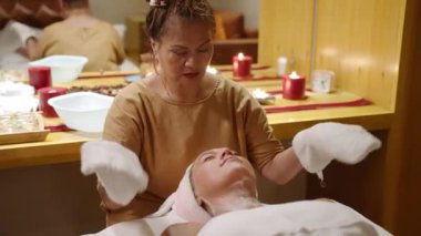 Asyalı kozmetikçi, sağlık merkezinde beyaz maskeyi kadın müşteri yüzünden siliyor. Çift ırklı güzellik uzmanı kaplıcada yüzünü sıcak beyaz havlularla temizleyen genç bir kadına yüz bakımı prosedürü uyguluyor.