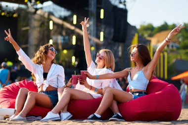 Sıradan giyinen mutlu kadınlar, kıyı etkinliğinde kutlama yaparlar. Üç genç bayandan oluşan bir grup, renkli fasulye koltukları ve açık hava müzik plaj festivalinde gün batımı ışıkları eşliğinde yaz havasının tadını çıkarıyor..