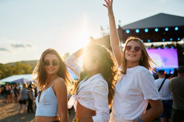 Kalabalık sahnenin yanındaki yaz atmosferinin tadını çıkarıyor. Güneşli plaj müzik festivalinde üç mutlu bayan arkadaş dans ediyor. Günlük giyinen kızlar kutlama yapar, gündüz vakti deniz kıyısında festival atmosferinde eğlenirler..