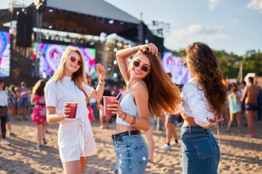 Mutlu dişiler kokteylleri sever, sahnenin yakınında kalabalık olur. Bir grup neşeli kız yaz güneşi altında plaj müzik festivalinde dans ediyor. Tropikal festival etkinliğinde arkadaşlık, parti yaşam tarzı.