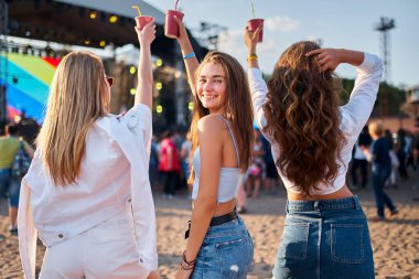 Kızlar arkaya bakıyor, canlı müzikten hoşlanıyor, yaz havası var. Bir grup genç kadın, güneşli plaj festivalinde soğuk içeceklerle tezahürat yapıyor. Arkadaşlar dans eder, kumlu sahilde içki bardaklarıyla kutlama yapar, sahneye bakar..