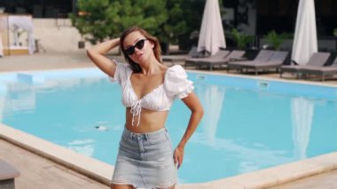 Lüks bir kulüpte etek, bluz ve güneş gözlükleriyle yüzme havuzunda poz veren ve gülümseyen güzel bir kız modeli. Yaz partisinde çekici bir kadın göletin berrak mavi sularının yanında tatil yapıyor..