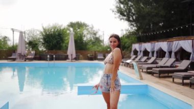 Güzel bir kadın kalıyor, poz veriyor, plaj restoranında yüzme havuzu ve güneşli yaz günlerinde arkadaşlarıyla flört ediyor. Mayolu ve şortlu bir model..