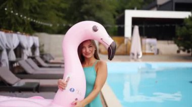 Mavi elbiseli, şişme flamingo tüplü, kameraya gülümseyen ve turkuaz yüzme havuzunda poz veren güzel bir kız. Güneşli ve güneşli bir tatil gününde güneşli plaj klübü.