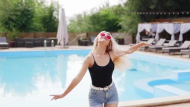 Sarı saçlı kadın kırmızı güneş gözlüğü, şort ve yüzme havuzunda güneş pansiyonları ve çam ağaçlarıyla fotoğraf çekmek için dans ediyor. Tatillerde poz vermek ve flört etmek.