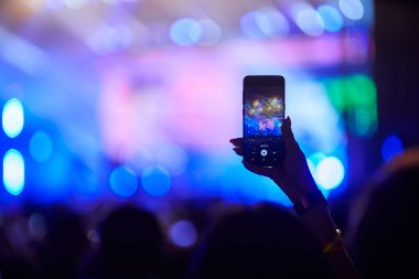 Enerjik festival atmosferi, hayranlar gece performansını seviyor. Konsere gidenler akıllı telefonlu canlı müzik etkinliği, kalabalığın üzerine kaldırılan el, sahne ışıklarını yakalayan, arka planda bulanık sahne sanatçıları..