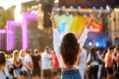 Barış işareti ile el kaldırdı, konserin tadını çıkarıyor. Yaz Sahili Müzik Festivali 'ndeki genç bayanın arka görüntüsü. Kalabalık, sahne ışıkları, günbatımı partileri. Sıradan moda, açık hava etkinliği, eğlenceli atmosfer..