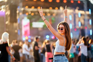 Canlı festival atmosferi, sahne ışıkları ile alacakaranlık ışığı. Mutlu genç kadın yaz müzik festivalini seviyor, barış tabelasıyla el kaldırıyor, plaj partisinde dans ediyor. Moda kalabalığı, tatil havası, eğlence..