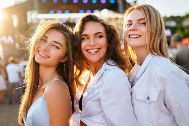 Arkadaşlar güneş ışığında poz verir, sahne ışıkları arkada. Üç gülümseyen genç kadın kumlu sahilde yaz müzik festivalinin tadını çıkarıyor. Açık hava konser etkinliğinde günlük moda, eğlence ve birliktelik..