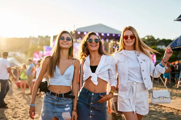 stock image Friends dance, enjoy live band, summer vibes by sea. Group of joyful young women at sunset beach music festival. Smiling girls with sunglasses, casual fashion at outdoor concert, vacation leisure.