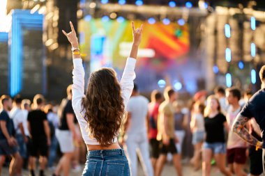 Happy crowd around stage, summer event vibe. Back view of young woman with raised hands enjoys live music at sunny beach festival. Fan cheers band, fun outdoor concert. Casual denim, festival fashion. clipart