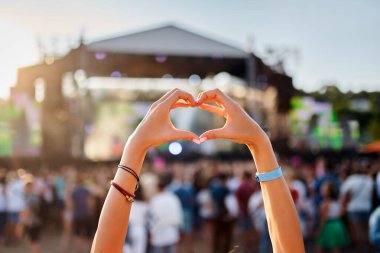 Konsere giden kişi kalp şeklini sahil kenarındaki müzik festivalinde elleri ile oluşturur, kalabalık günbatımında canlı performansı sever, yaz etkinliğinde aşk sembolü jesti, açık hava sanatının gençlik kutlamaları.