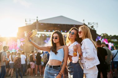 Modayı takip eden yaz kıyafetleri içindeki kızlar konser ve parti atmosferinden hoşlanırlar. Bir grup mutlu genç bayan arkadaş, güneşli plaj müzik festivalinde selfie çekiyor. Gençlik kültürü, arkadaşlık, açık hava eğlencesi.