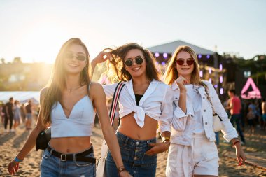 Arkadaşlar gülüyor, güneşin altında dans ediyorlar. Yazın plaj müzik festivalindeki kızlar. Genç kadınlar festival atmosferini seviyor, son moda güneş gözlüğü takıyor, günlük kıyafetler giyiyor. Partiye katılanlar açık hava etkinliğini birlikte kutluyorlar.