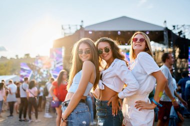 Arkadaşlar dans eder, güneşte güler, deniz kenarında. Üç genç bayandan oluşan bir grup plajda yaz müzik festivalinin tadını çıkarıyor. Arka planda kalabalık olan parti atmosferi. Günlük kıyafet, güneş gözlüğü, altın saat eğlencesi..