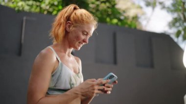 Gülümseyerek sağlık verilerini izliyor ve huzurlu bir ortamda ideal eğitim sonuçlarını sağlıyor. Kızıl saçlı bayan sporcu, şık bir açık hava tropikal spor salonunda spor sonrası akıllı telefon uygulamasını kontrol ediyor. Slomo