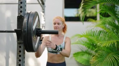 Odaklanmış bayan sporcu halter ekipmanları hazırlıyor, güvenliği sağlıyor. Atletik kadın açık hava spor salonunda halterin çene kilidini ayarlıyor. Forma giymiş kızıl saçlı ağırlık kaldırıyor, dayanıklılığını gösteriyor..