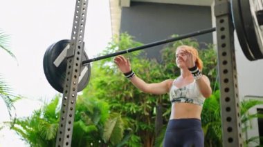 Kaslı kadın bacak gücünü çalıştırıyor, halter kaldırma zorluğuyla karşı karşıya olduğu belli. Kızıl saçlı atletik kadın, çömelmek için ağır halterleri kaldırıyor. Açık hava spor salonu, tropik bitkilerle çevrili. Yavaşla