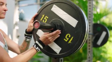 Kadın ağırlık plakalarını güvenli taşıma antrenmanı ve güç antrenmanı için ayarlıyor. Spor meraklısı, güneşli açık hava spor salonunda halterdeki çene kilidini emniyete alıyor. Eller ekipmanları hazırlasın, güvenli çalışma oturumu sağlansın.
