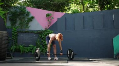 Fitness meraklısı halter kaldırır, güç ve beceri gösterir. Kaslı kadın açık hava spor salonunda çömelme hareketleri yapıyor. Güneş ışığıyla aydınlanan sahne, güçlendirme, kırsal spor salonundaki adanmışlık. Yavaşla