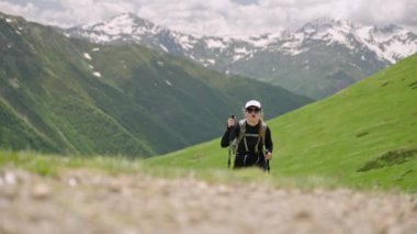 Kadın sırt çantasıyla Svaneti dağlarına tırmanıyor. Yeşil yamaçlarda yürüyüş yapan yürüyüşçüler, karlı zirveler. Açık hava macerası, dağ yürüyüşü, yürüyüş direkleri fiziksel aktivite. Yavaş çekim.